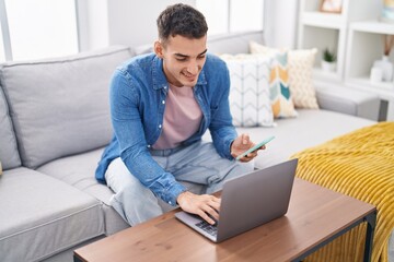 Young hispanic man using laptop and smartphone sitting on sofa at home