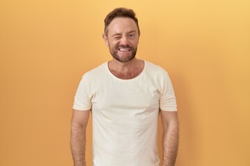 Middle age man with beard standing over yellow background winking looking at the camera with sexy expression, cheerful and happy face.