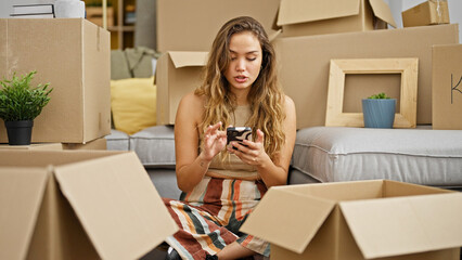 Young beautiful hispanic woman using smartphone sitting on floor at new home