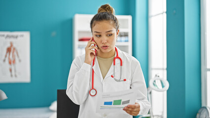 Young beautiful hispanic woman doctor talking on smartphone with relaxed expression reading medical report at clinic