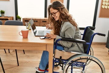 Young beautiful hispanic woman business worker writing on document sitting on wheelchair at office