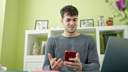 Young hispanic man using smartphone and laptop looking upset at dinning room