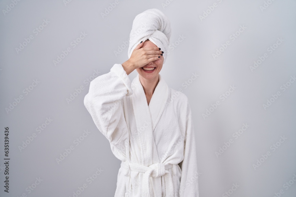 Poster blonde caucasian woman wearing bathrobe smiling and laughing with hand on face covering eyes for sur