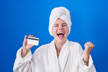 Blonde caucasian woman wearing bathrobe holding credit card screaming proud, celebrating victory and success very excited with raised arms