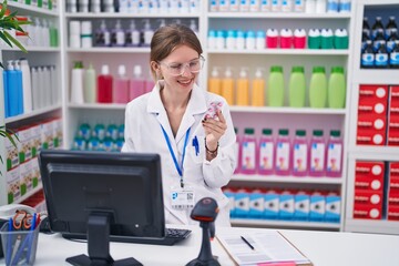 Young blonde woman pharmacist using computer holding pills smiling at pharmacy