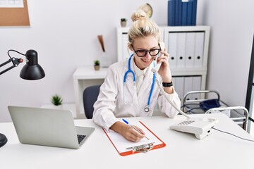 Young blonde woman doctor talking on telephone writing on document at clinic