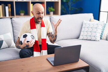 Hispanic man with tattoos watching football match hooligan holding ball on the laptop celebrating...