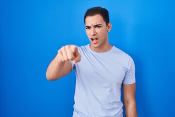 Young hispanic man standing over blue background pointing displeased and frustrated to the camera,...