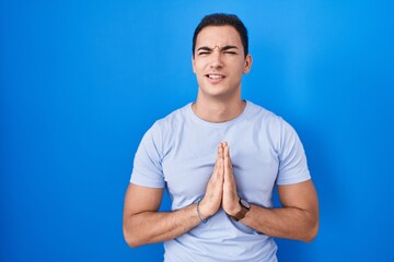 Young hispanic man standing over blue background begging and praying with hands together with hope...