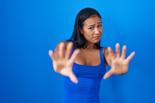 Hispanic woman standing over blue background afraid and terrified with fear expression stop gesture with hands, shouting in shock. panic concept.