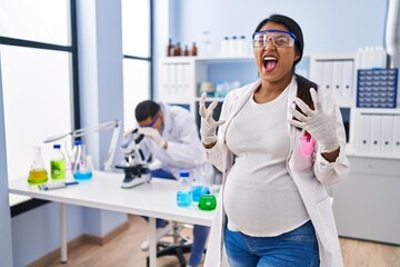Young hispanic woman expecting a baby working at scientist laboratory crazy and mad shouting and...