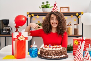 Hispanic woman with curly hair celebrating birthday holding big chocolate cake pointing finger to one self smiling happy and proud