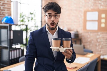 Hispanic man with beard working at the office holding coffee scared and amazed with open mouth for surprise, disbelief face