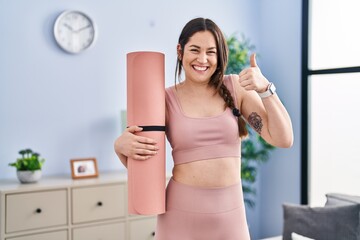 Fototapeta na wymiar Young brunette woman holding yoga mat smiling happy and positive, thumb up doing excellent and approval sign