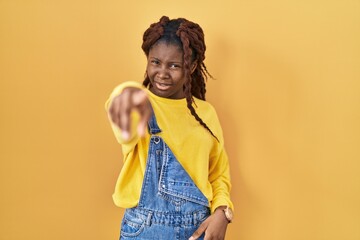 African woman standing over yellow background pointing displeased and frustrated to the camera,...