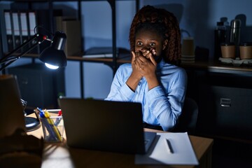 Naklejka na ściany i meble African woman working at the office at night shocked covering mouth with hands for mistake. secret concept.