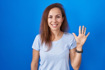 Brunette woman standing over blue background showing and pointing up with fingers number five while smiling confident and happy.