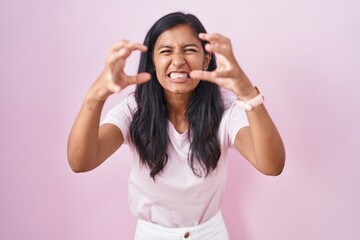 Young hispanic woman standing over pink background shouting frustrated with rage, hands trying to strangle, yelling mad