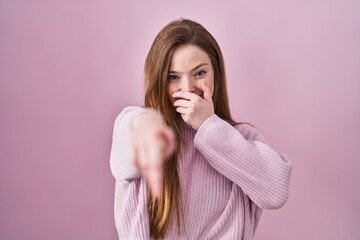 Young caucasian woman standing over pink background laughing at you, pointing finger to the camera...