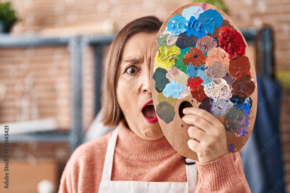 Poster middle age hispanic woman holding painter palette close to face in shock face, looking skeptical and