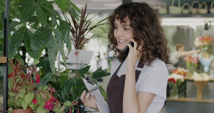 Young lady in apron talking on mobile phone and laughing standing in flower shop looking at green plants. Floristry business and communication concept.