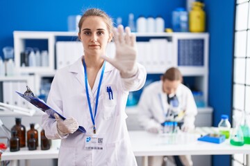 Blonde woman working at scientist laboratory with open hand doing stop sign with serious and confident expression, defense gesture