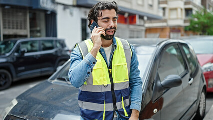 Young hispanic man standing by car wearing reflective vest talking on smartphone at street