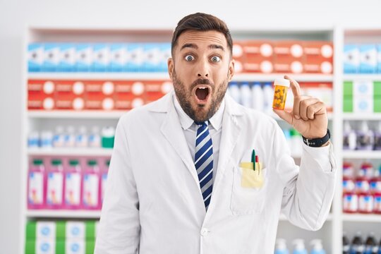 Handsome hispanic man working at pharmacy drugstore holding pills scared and amazed with open mouth for surprise, disbelief face