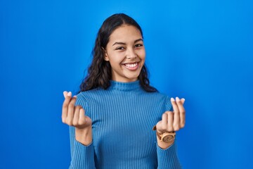Young brazilian woman standing over blue isolated background doing money gesture with hands, asking...