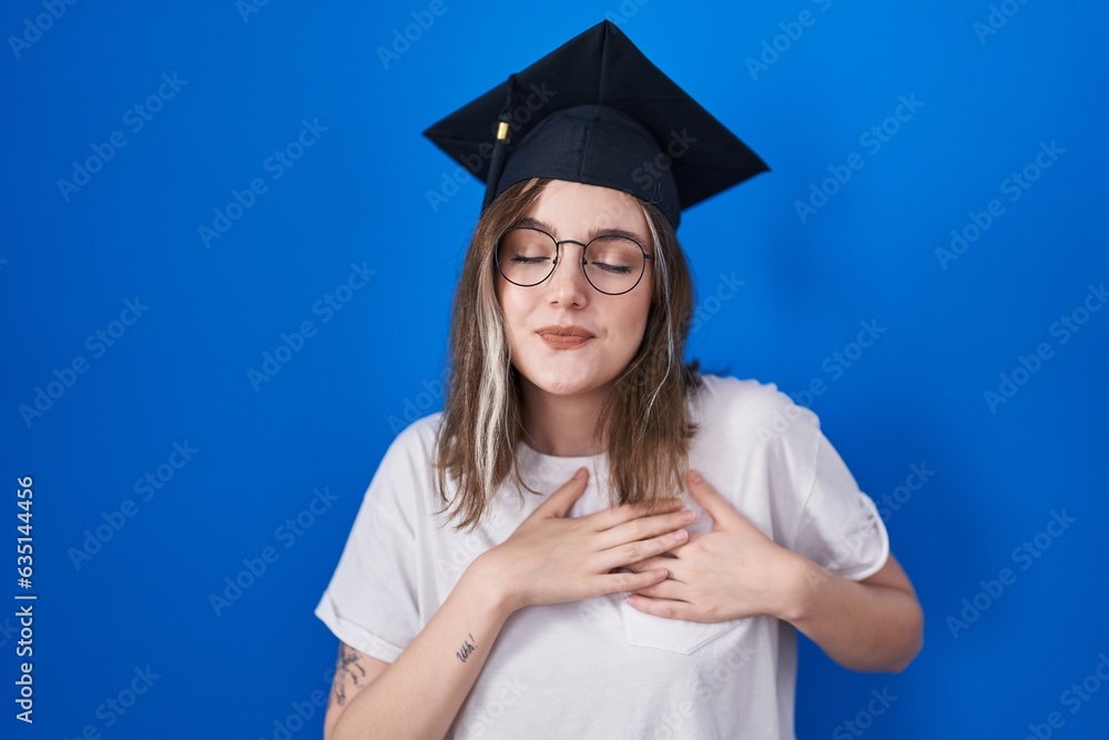 Poster blonde caucasian woman wearing graduation cap smiling with hands on chest with closed eyes and grate