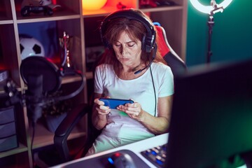 Middle age woman streamer playing video game using smartphone at gaming room