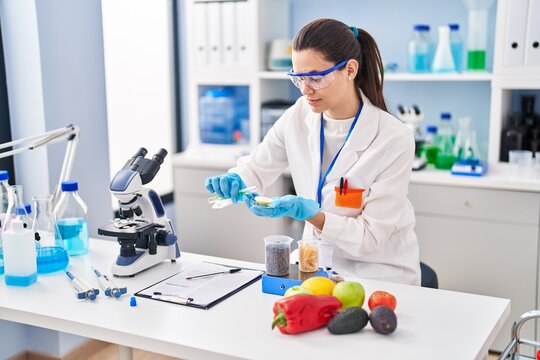 Young beautiful hispanic woman scientist pouring liquid on piece of apple at laboratory