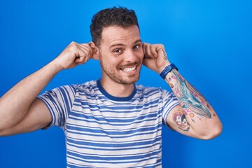 Young hispanic man standing over blue background smiling pulling ears with fingers, funny gesture....