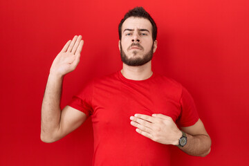 Young hispanic man wearing casual red t shirt swearing with hand on chest and open palm, making a...