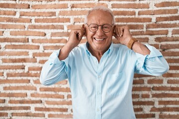 Senior man with grey hair standing over bricks wall smiling pulling ears with fingers, funny...