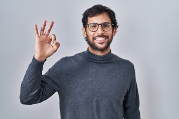 Fototapeta premium Handsome latin man standing over isolated background smiling positive doing ok sign with hand and fingers. successful expression.