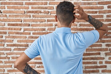Brazilian young man standing over brick wall backwards thinking about doubt with hand on head