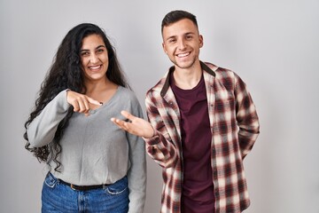 Young hispanic couple standing over white background amazed and smiling to the camera while presenting with hand and pointing with finger.