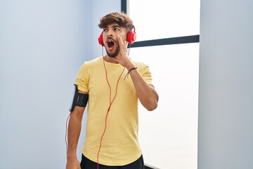 Arab man with beard wearing sportswear and headphones in shock face, looking skeptical and...