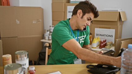 Young hispanic man volunteer using touchpad talking on smartphone checking products at charity center