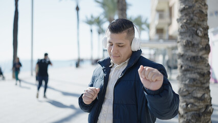 Young hispanic man listening to music and dancing at seaside