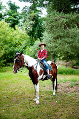 little handsome blonde smiling boy in red checkered shirt and cowboy brown hat riding horse in green forest on sunny day	