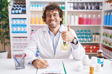 Hispanic young man working at pharmacy drugstore doing happy thumbs up gesture with hand. approving expression looking at the camera showing success.