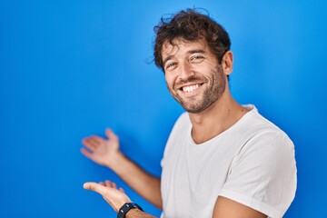 Hispanic young man standing over blue background inviting to enter smiling natural with open hand