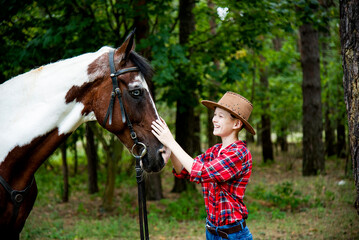 Obraz premium little handsome blonde smiling boy in red checkered shirt and cowboy brown hat walking and hugging horse in green forest on sunny day 