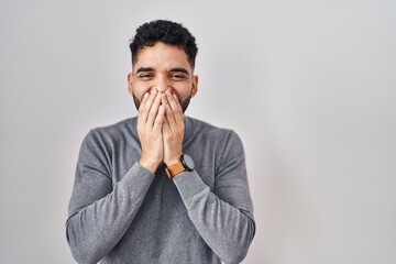 Hispanic man with beard standing over white background laughing and embarrassed giggle covering...