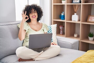 Young brunette woman with curly hair using laptop sitting on the sofa at home smiling positive...