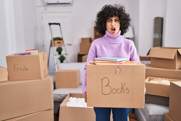 Young brunette woman with curly hair moving to a new home holding cardboard box angry and mad screaming frustrated and furious, shouting with anger. rage and aggressive concept.