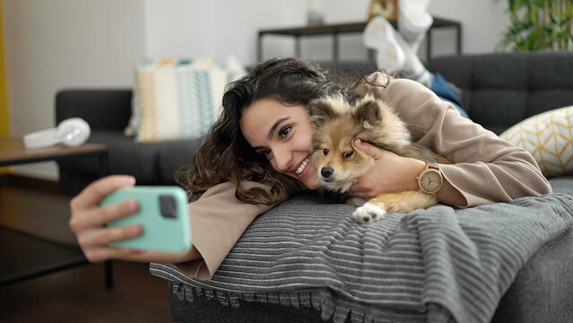 Young hispanic woman with dog taking selfie picture with smartphone lying on sofa at home