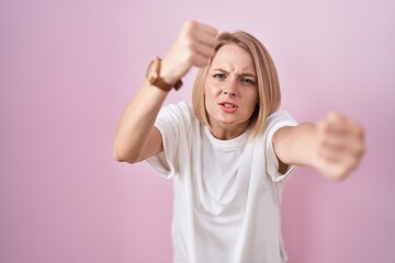 Young caucasian woman standing over pink background angry and mad raising fists frustrated and furious while shouting with anger. rage and aggressive concept.